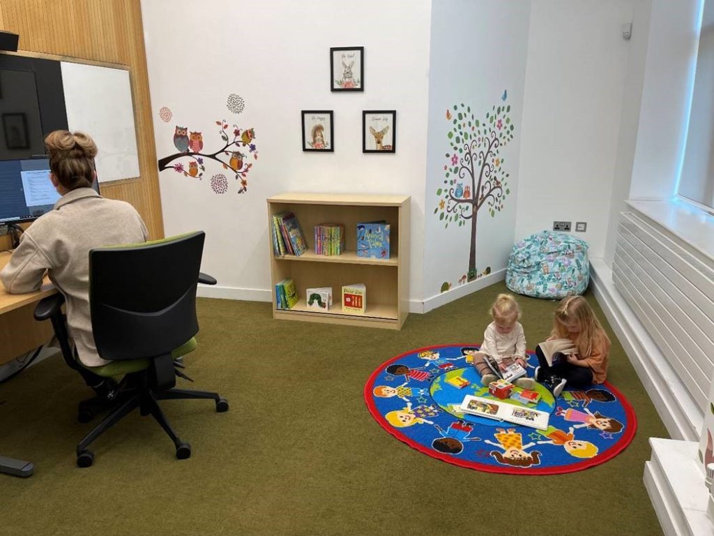 The photograph shows the Library's new Family Room. The room has a desk and computer for work, while the floor has lots of child-friendly resources. There are decorations on the wall and a mat on the floor to make the space more welcoming for our little visitors. 
