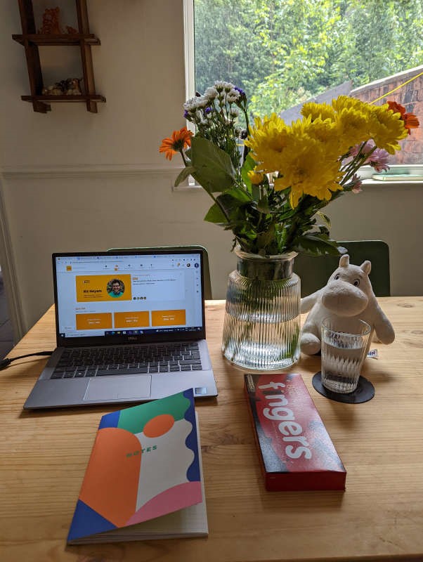 A large wooden table with a laptop on it. There is a large vase of flowers to the right, and a Moomin soft toy sits beneath the vase. 